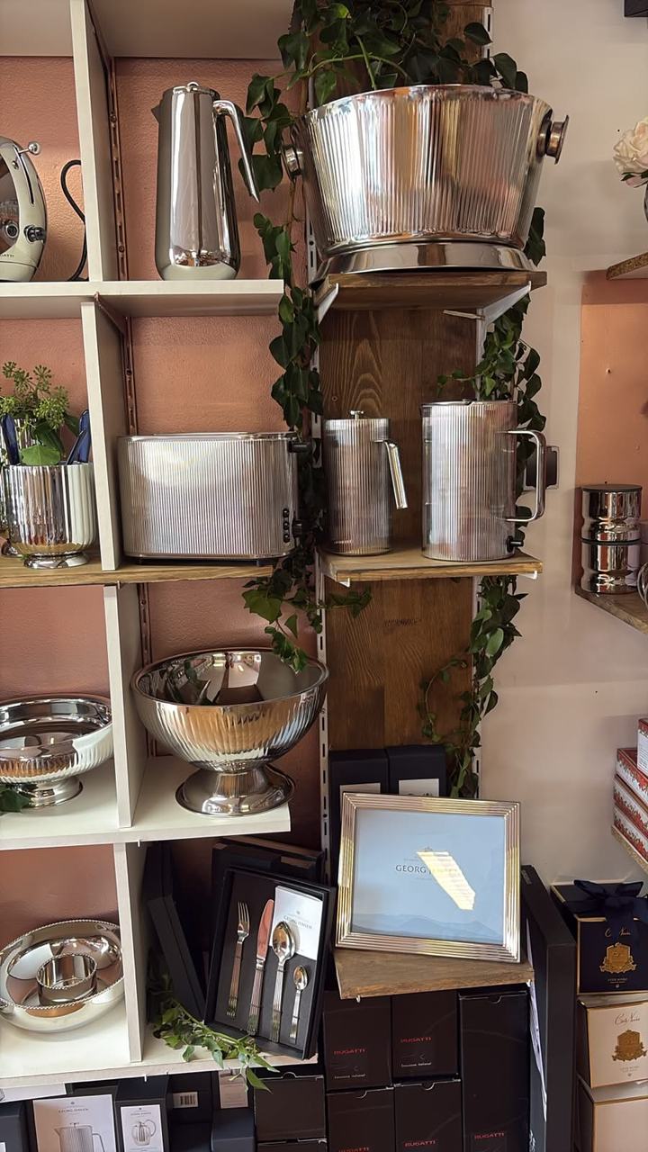 Un mur de la boutique : seau à champagne et pièces en acier cannelé entre le lierre, coffret de couverts Georg Jensen au pied des étagères.